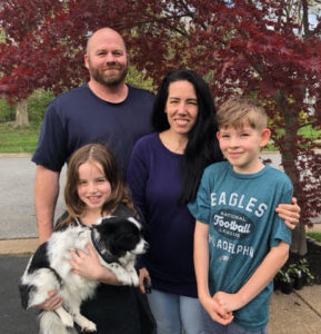 Larisa forest with her husband, son and daughter, who is holding the dog are standing outside in front of a Japanese maple tree.
