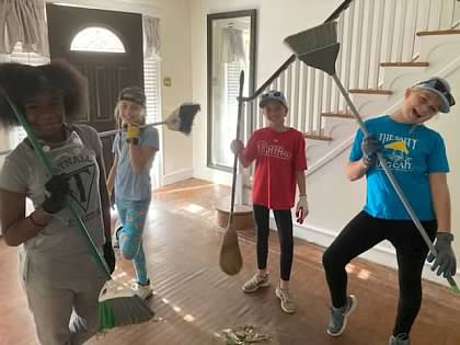 Four children as posing for the camera while holding brooms. There is a pile of dirt on the floor in front of them.